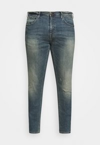 Vaqueros azul deslavado con un ajuste slim, que cuentan con cinco bolsillos, ligero desgaste y un cierre de botón de metal. La textura del denim es visible.