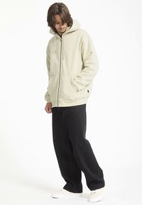 Sweat à capuche beige clair avec fermeture éclair, capuche doublée, manches longues et poches avant, associé à un pantalon noir à jambes larges et des chaussures blanches.