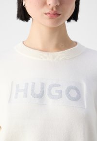 Personne aux cheveux courts et foncés portant un pull blanc avec « HUGO » en lettres argentées sur la poitrine et une boucle d'oreille anneau argenté.