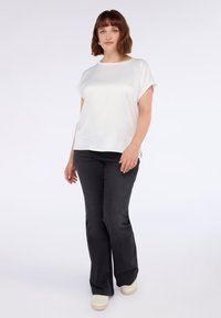 Femme aux cheveux bruns courts portant un haut blanc à manches courtes, un jean noir et des baskets blanches, se tenant devant un fond clair uni.