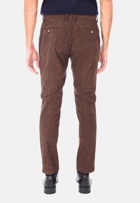 Uomo che indossa pantaloni di velluto a coste marroni e scarpe nere, visto da dietro su uno sfondo chiaro e uniforme.
