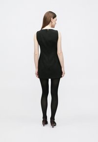 Vestido mini preto sem mangas com silhueta justa, apresentando um discreto fecho de correr nas costas e combinado com collants pretos e sapatos de salto elegantes.