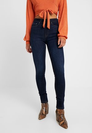 Vrouw draagt donkerblauwe skinny jeans, een oranje crop top met lange mouwen en een strik in de taille, en bruine enkellaarsjes met een slangenhuidpatroon.