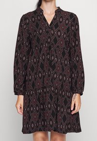 Svart klänning med ett geometriskt diamantmönster i rött och vitt. Har långa ärmar, V-hals och en utsvängd kjoldesign.