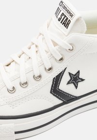 Sneaker alta in tela bianca con logo a stella nera, occhielli metallici, lacci bianchi e dettaglio della suola in gomma nera. Presenta cuciture a contrasto.