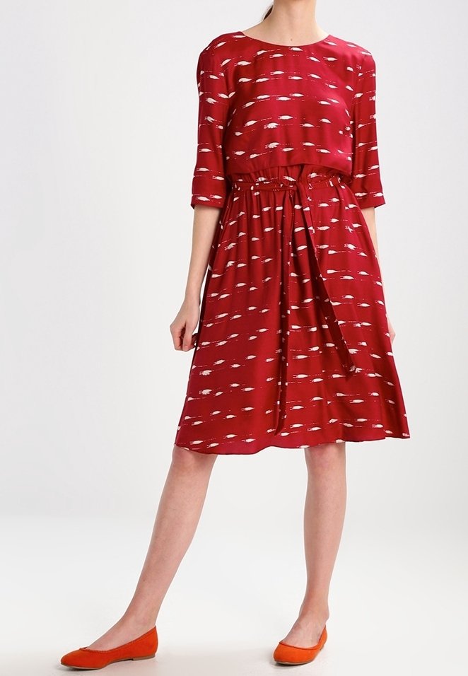 Femme portant une robe rouge mi-longue avec des motifs blancs, des manches trois-quarts, une ceinture à la taille et des chaussures plates orange.