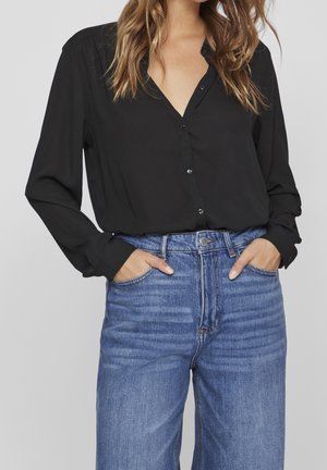 Vrouw draagt een zwarte blouse met lange mouwen en knopen, ingestopt in een high-waist blauwe jeans, met handen in de voorzakken.