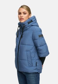 Naine sinises lühikestes varrukates pufferjacketis, millel on kõrge krae ja mustad nöörid, kannab tumedaid teksaseid lihtsa tausta ees.