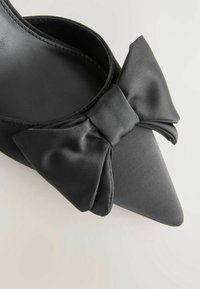 Chaussure plate noire à bout pointu avec grand nœud en satin sur le devant, doublure intérieure visible, sur un fond clair et neutre.