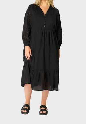 Vrouw draagt een zwarte midi-jurk met lange mouwen, knoopdetails en kanten mouwen, gecombineerd met zwarte gevlochten sandalen op een effen achtergrond.