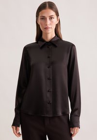 Donna che indossa una camicia nera in satin con maniche lunghe e bottoni, con colletto, in piedi contro uno sfondo beige chiaro e uniforme.