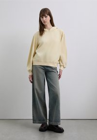 Gelber übergroßer Hoodie, weit geschnittene ausgewaschene Jeans und klobige schwarze Stiefel. Glatter Stoff, lässiges Design und entspannte Passform. Minimalistischer Hintergrund.