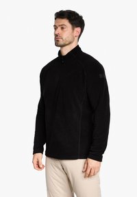 Schwarzer Fleece-Pullover mit hohem Kragen, halbem Reißverschluss und strukturiertem Stoff. Mit kontrastierenden Nähten und einem Logo am Ärmel.