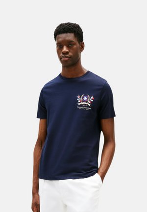 Giovane uomo con capelli corti e ricci indossa una maglietta blu navy con logo stemma multicolore e pantaloni bianchi, in piedi con una mano in tasca.