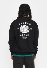 Zwarte sweatshirt met een witte grafiek van een vintage espressomachine op de achterkant, met de tekst "SACRED ALTAR 1995" en aanvullende zinnen.