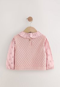 Blouse rose pour bébé avec broderie florale ajourée, col volanté et surcouche sans manches tricotée, suspendue sur un cintre en bois sur fond blanc.