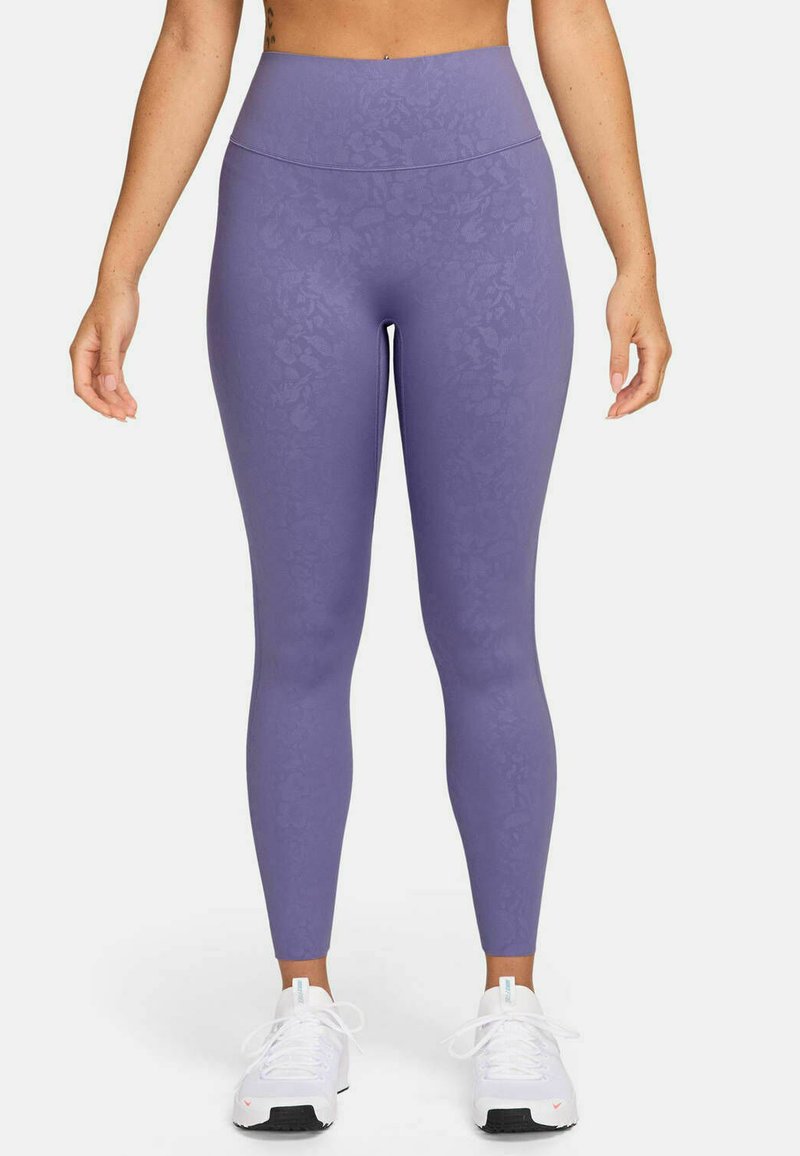Lila leggings med ett texturerat blommönster. Hög midja; slät, elastisk tyg. Bärs med vita träningsskor.