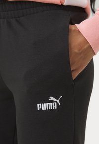 Črne bombažne trenirke s stransko žepom, ki imajo bel logo Puma. Tkanina je gladka in ima elastičen pas.