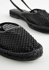 Svart nät espadrillo med pärldekorationer, platt sula, textilöverdel och vävt jute-trim. Har justerbara remmar för en säker passform.