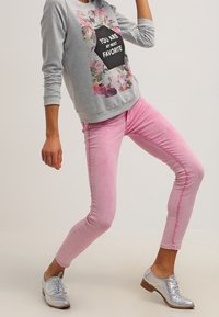 Henkilö, joka on pukeutunut harmaaseen huppariin, jossa on kukkakuosi ja teksti, vaaleanpunaisiin tiukkoihin farkkuihin ja kiiltäviin hopeisiin nauhakengiin dynaamisessa asennossa.