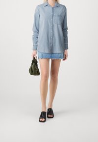 Woman wearing light blue denim shirt, frayed denim mini skirt, black open-toe mules, holding small olive green handbag.