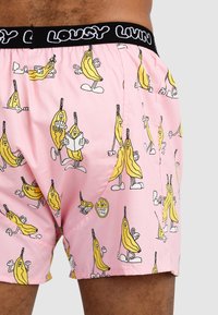 Pink boxer shorts with a banana print in black and yellow, featuring a black waistband with "LOUSY LIVIN" text, made from lightweight fabric.