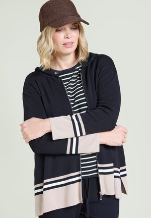 Woman wearing a brown cap and black zip-up sweater with beige and white stripes, standing with arms crossed and looking down.