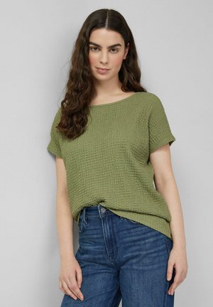 Woman with long wavy hair wears textured olive green short-sleeve top and blue jeans, standing against a plain light gray background.