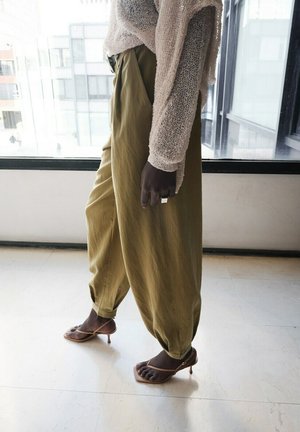 Personne debout à l'intérieur portant un pantalon ample olive, un pull beige transparent, des sandales à talons avec brides à bout carré, et une grosse bague argentée à la main.