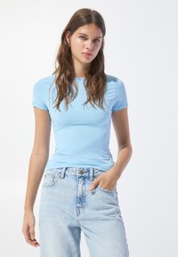 T-shirt aderente azzurro chiaro, realizzata in tessuto morbido, con maniche corte e scollo rotondo, abbinata a jeans chiari a vita alta.