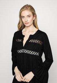 Vestido negro de manga larga con escote en V; presenta recortes de encaje en forma circular y un diseño en capas con mangas acampanadas. Textura de tela suave.