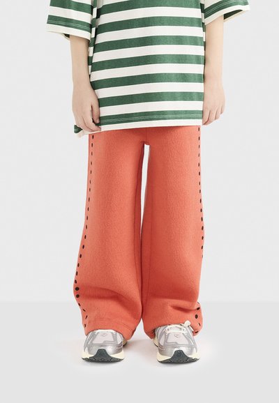 Breddebenede, højtaljede bukser i tekstureret orange stof med sort sidesyning. Kombineret med en overdimensioneret t-shirt med grønne og hvide striber og sneakers.