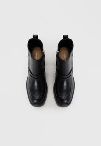 Bottines en cuir noir avec une texture lisse, dotées d'une bride sur le devant, d'une fermeture éclair sur le côté et d'une semelle plate striée.