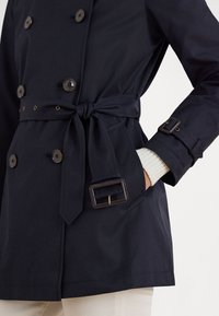Tmavomodrý dvojradový trenčkot s opaskom, vybavený hnedými gombíkmi, zlatým prackovým detailom a vnútorným vreckom s rebrovaným manžetou.