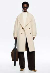 Cappotto in lana avorio doppiopetto con ampi rever, tasche frontali e pantaloni marroni a contrasto. Gli occhiali da sole neri completano il look.