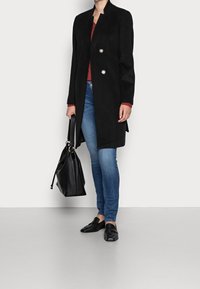 Frau trägt einen schwarzen langen Mantel, ein rotes Oberteil, blaue Jeans und schwarze Slipper, hält eine große schwarze Handtasche vor einem schlichten hellen Hintergrund.