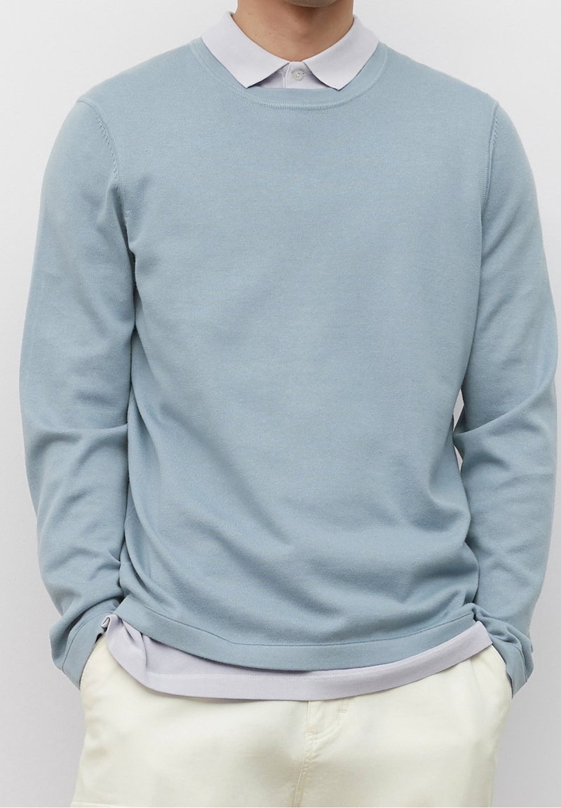 Pullover azzurro chiaro realizzato in materiale morbido, con maniche lunghe e scollatura rotonda, indossato sopra una camicia colletto grigia chiara.