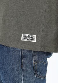 Grå t-shirt i mjukt tyg med rak passform, med en vit etikett som står "DENIM FACTORY" på fållen. Matchas med blå denimjeans.