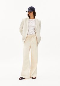 Blazer beige, camicia a righe, pantaloni beige a gamba larga e berretto blu navy. Sandali con suola scura. Design minimalista con colori neutri.