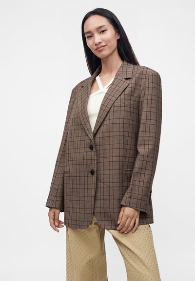 Femme aux longs cheveux foncés portant un blazer oversize à carreaux marron, un haut blanc et un pantalon beige texturé, debout devant un fond uni.