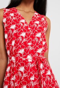 Abito rosso floreale con motivi a fiori bianchi, scollo a V, senza maniche e chiusura con bottoni. Vita con lacci per un look aderente.