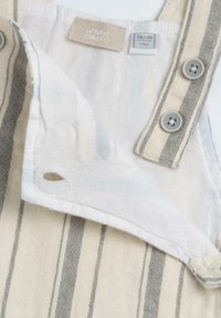 Salopette en tissu couleur crème avec des rayures grises, dotée d'une étiquette, de deux fermetures à boutons et d'une petite ouverture sur le côté.