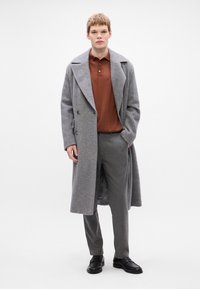 Grå hørfanget overfrakke med store revers, dobbeltbrystet foran, båret over en brun polo skjorte og grå bukser, parret med sorte sko.