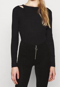 Mustat ribbed pitkähihaiset paita, jossa on leikattu olkapääyksityiskohta, yhdistettyinä korkeavyötäröisiin mustiin corduroy-housuihin, joissa on vetoketjukiinnitys ja metallirengasaksentti.