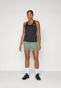 Melns tanktops ar mazu logotipu, zaļas šorti ar fakturētu rakstu, balti zeķi un melni sporta apavi uz neitrāla fona.