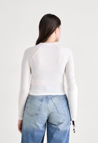 Weißes Langarmoberteil mit tailliertem Design und Raglanärmeln, kombiniert mit hellblauen hochgeschnittenen Jeans. Glatte Textur; minimalistischer Stil.