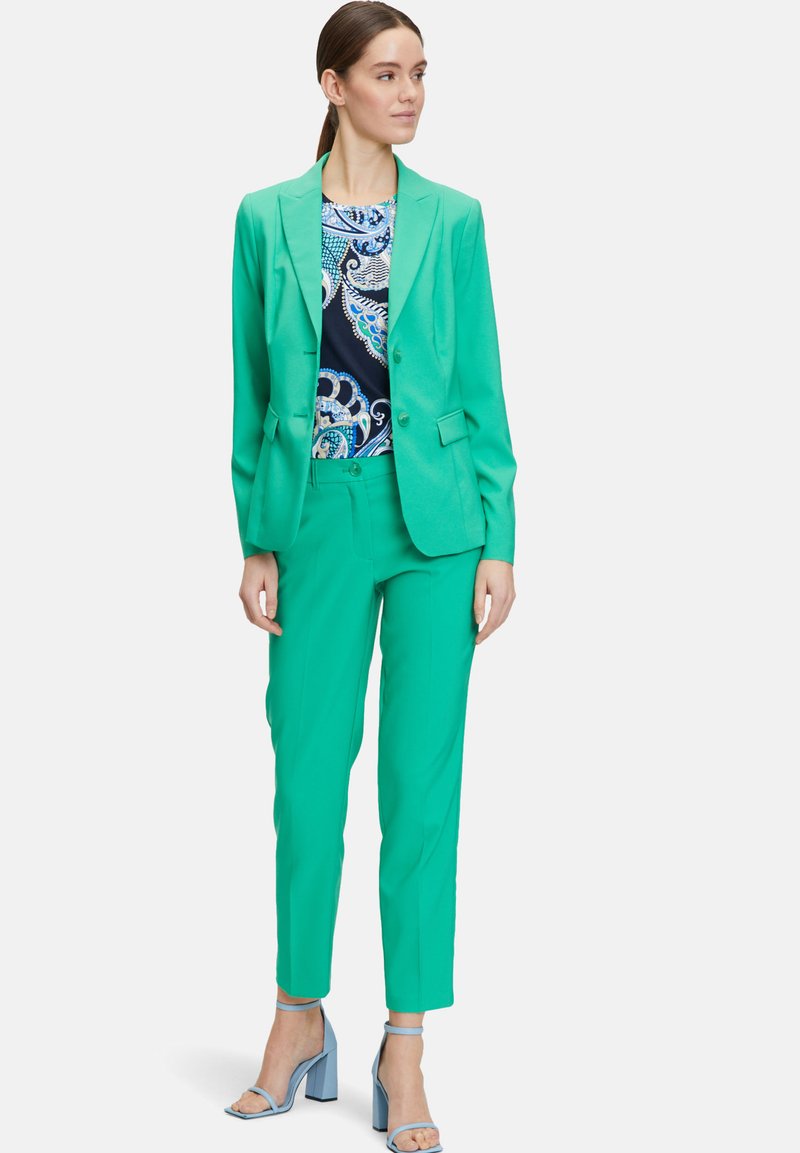 Světlý zelený oblek s upraveným blazerem a slim-fit kalhotami, doplněný vzorovaným topem a zakončený světle modrými sandály na blokovém podpatku.