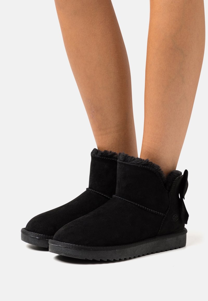 Bottines noires en daim avec un bord en fausse fourrure et une semelle plate en caoutchouc. Elles présentent un nœud décoratif à l'arrière et des détails de coutures contrastantes.