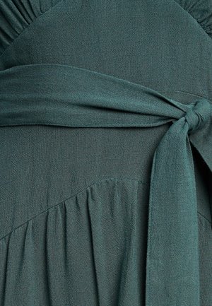 Tamnozelena haljina od mekog, teksturiranog materijala. Ima naborani izrez, pojas oko struka i rašireni kroj s suptilnim naborima.