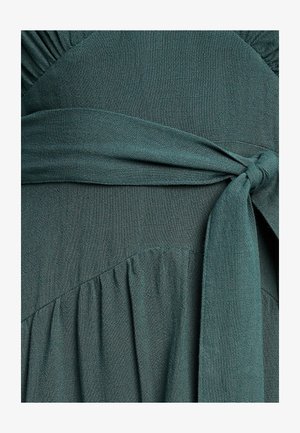 Tamnozelena haljina od mekog, teksturiranog materijala. Ima naborani izrez, pojas oko struka i rašireni kroj s suptilnim naborima.